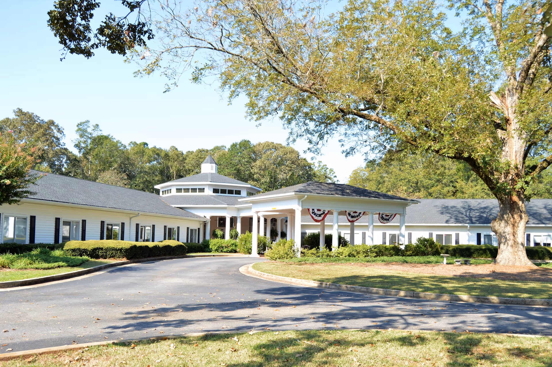 Exterior view of Mulberry Grove Senior Living facility showing a single-story white building with black shutters, a circular driveway, and a covered entrance decorated with patriotic bunting. Large trees and greenery surround the building under a clear blue sky.