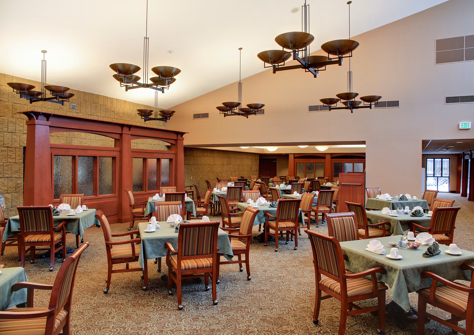 A spacious dining room with multiple tables covered in green tablecloths, each set with white napkins, cups, and saucers. The room features wooden chairs with striped cushions, large decorative light fixtures hanging from the ceiling, and a beige carpet with a subtle pattern. There is a wooden partition with frosted glass panels along one wall and an exit door visible in the background.