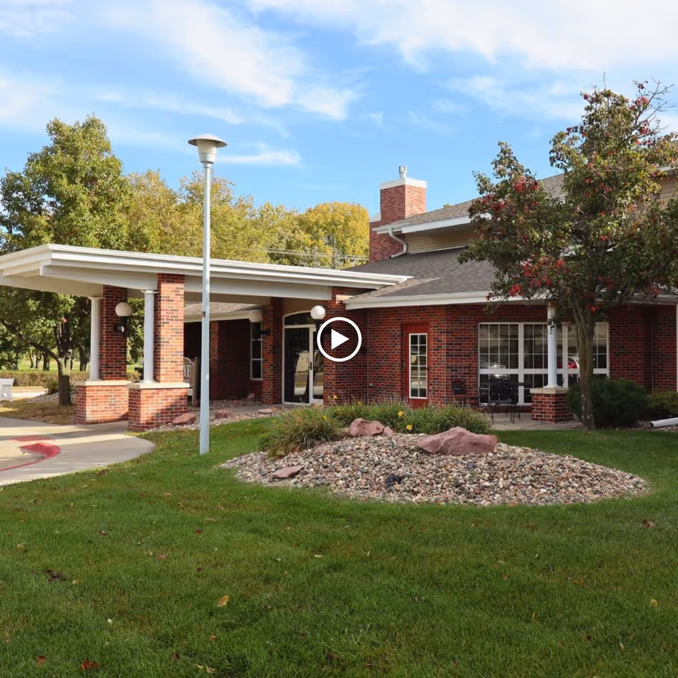 Exterior view of a brick building with a covered entrance, surrounded by green grass, trees, and a landscaped rock garden under a blue sky with some clouds.