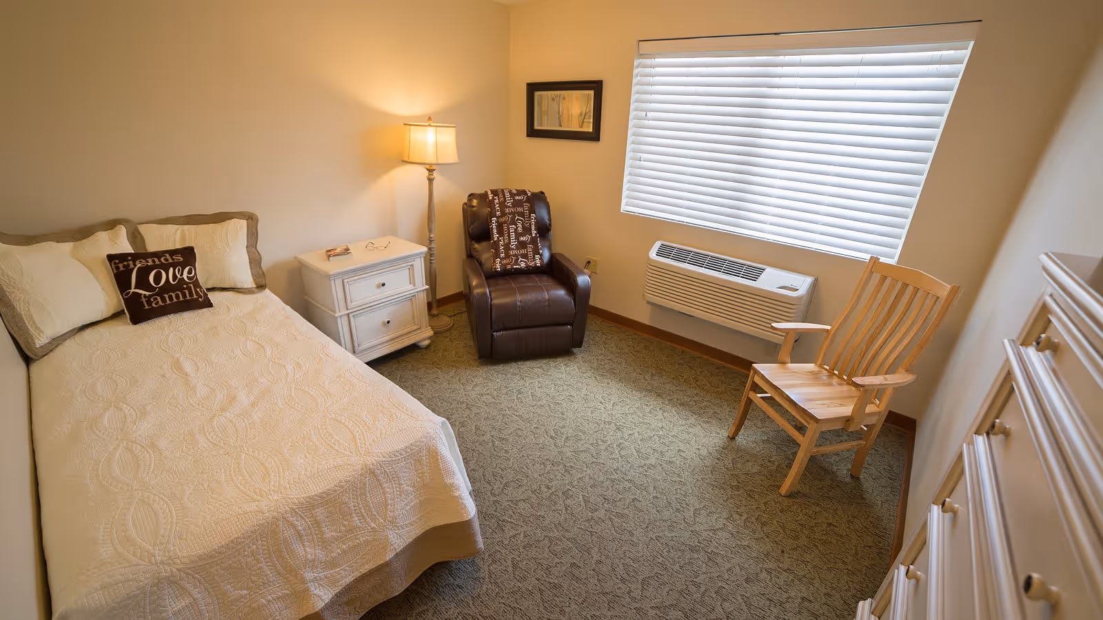 A cozy bedroom with a single bed covered in a cream-colored quilt and pillows, including one with the words 'friends Love family'. Next to the bed is a white nightstand with a lamp and a pair of glasses on it. There is a brown leather recliner chair with a decorative pillow, a wooden rocking chair, a window with closed blinds, and a wall-mounted air conditioning unit beneath the window. The room has beige walls and carpeted flooring.