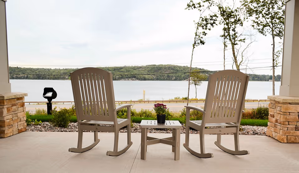 Two beige rocking chairs with armrests facing a large body of water, separated by a small matching table with a potted plant on top, on a covered patio with stone pillars and a view of trees and hills across the water.