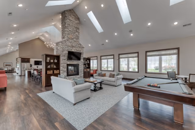 Spacious and well-lit common area with a stone fireplace at the center, two sofas facing each other on a large rug, a pool table on the right, and large windows letting in natural light. The room has high vaulted ceilings with skylights and wooden flooring, with a kitchen and dining area visible in the background.
