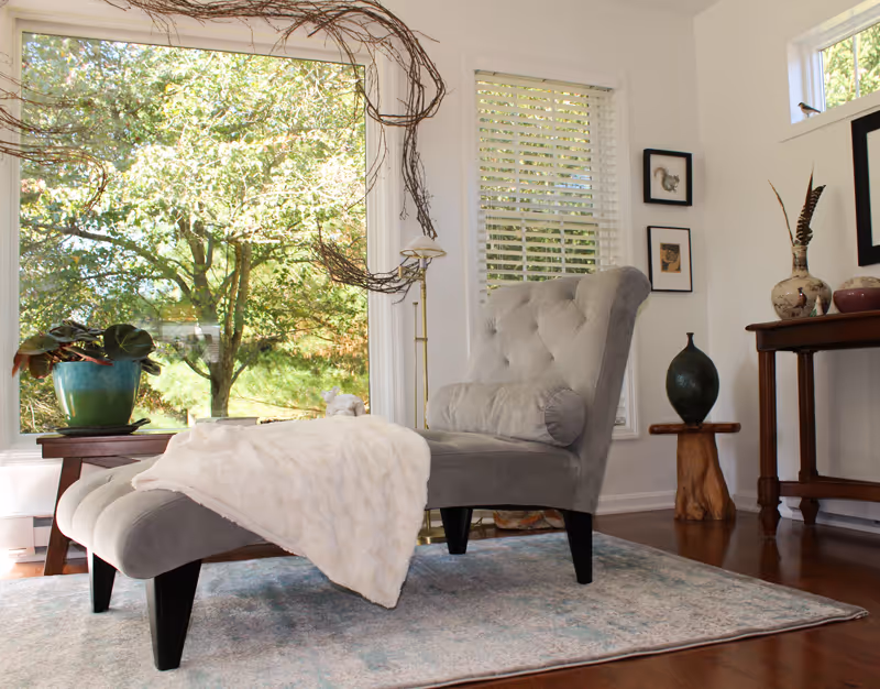 A cozy living room corner with a gray tufted armchair and matching ottoman draped with a white blanket, placed on a light blue patterned rug. Large windows behind the chair show green trees outside. A wooden side table with a green potted plant and a wooden console table with decorative vases and framed artwork on the wall are also visible.