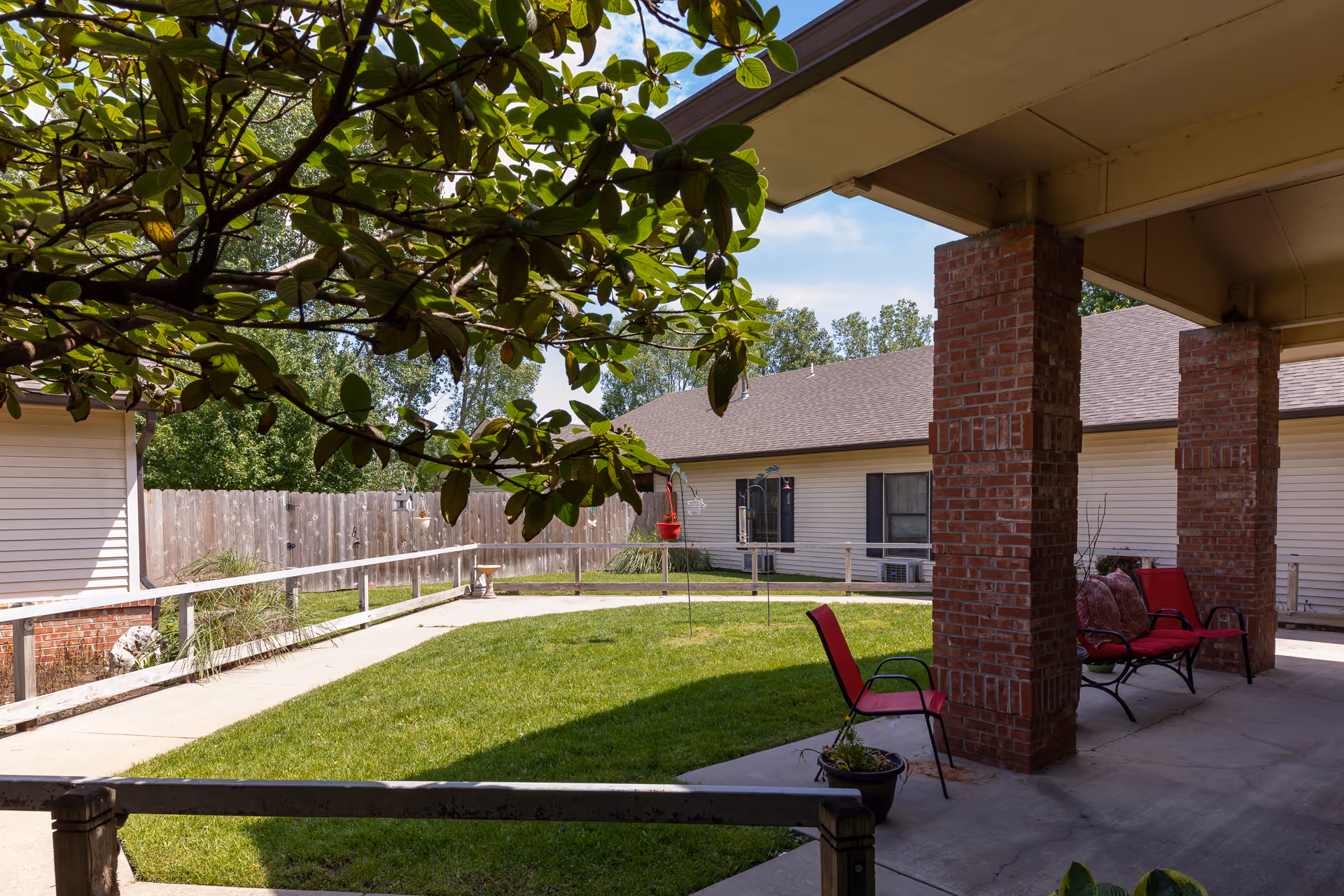 Outdoor patio area at Homestead Senior Living of El Dorado with red cushioned chairs under a covered porch supported by brick pillars, a green lawn, a concrete walkway with handrails, and a wooden fence in the background under a partly cloudy sky.