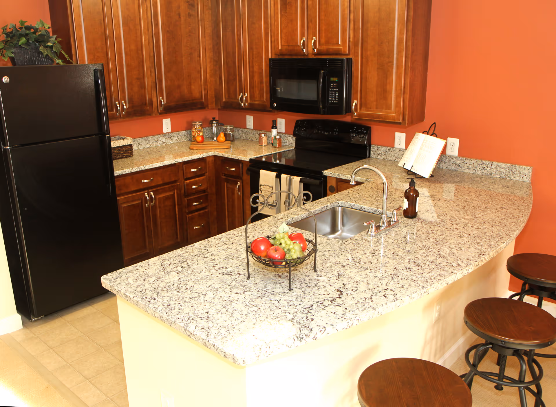 A kitchen area with granite countertops, a double sink, a black refrigerator, a black microwave above a black stove, wooden cabinets, and three wooden bar stools. There is a fruit basket with apples and grapes on the counter, a soap dispenser near the sink, and a cookbook on a stand against a reddish-orange wall.