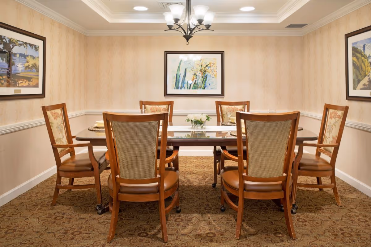 A dining room with a rectangular wooden table surrounded by six wooden chairs with cushioned seats and backs. The room has beige patterned wallpaper, a carpeted floor with a floral design, and three framed paintings on the walls. A chandelier with five lights hangs from the ceiling above the table, which is set with placemats and a small floral centerpiece.
