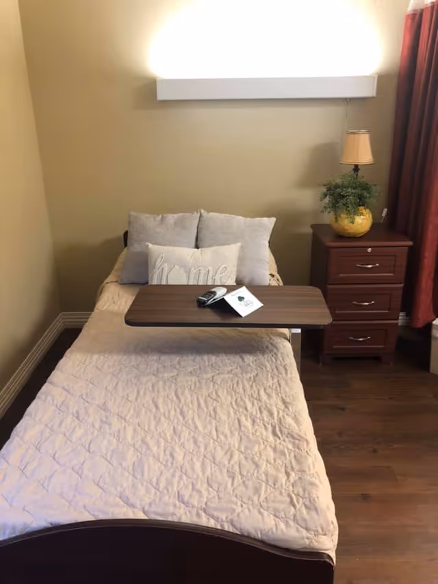 A single bed with beige quilt and three pillows, one of which has the word 'home' embroidered on it. A wooden overbed table holds a TV remote and a folded paper. Next to the bed is a wooden nightstand with three drawers, a yellow vase with green plant, and a small lamp. The room has beige walls, wooden floor, and red curtains on the right side.