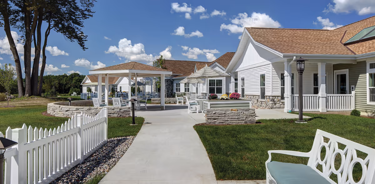 Outdoor view of Cedar Community Memory Care - The Cottages featuring a paved walkway, white picket fence, white benches, a gazebo, patio umbrellas, and a building with white siding and stone accents under a blue sky with scattered clouds.