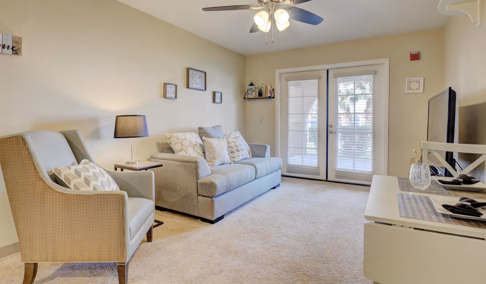 A cozy living room with a gray sofa adorned with patterned pillows, a cushioned armchair with a decorative pillow, a side table with a lamp, a ceiling fan with lights, and a TV on a white stand. French doors with blinds lead to an outdoor area, and the walls are decorated with small framed pictures.