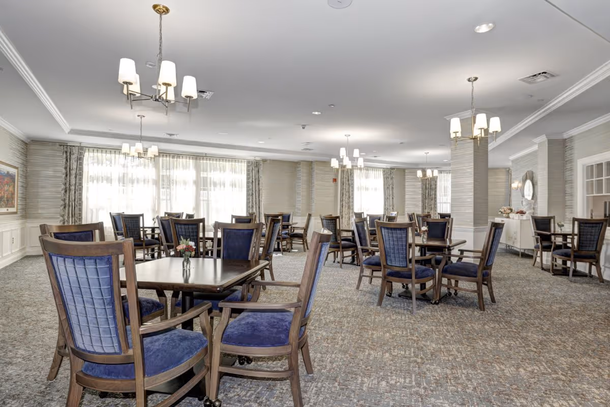 Spacious dining room with multiple dark wood tables and blue upholstered chairs beneath chandeliers and large curtained windows.