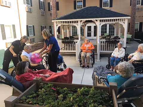 A group of elderly people, some in wheelchairs and others seated, are gathered outdoors in a courtyard area of a senior living facility. Two caregivers are assisting and interacting with the residents. The courtyard features a gazebo structure and a raised garden bed with plants in the foreground. The building of the facility is visible in the background.