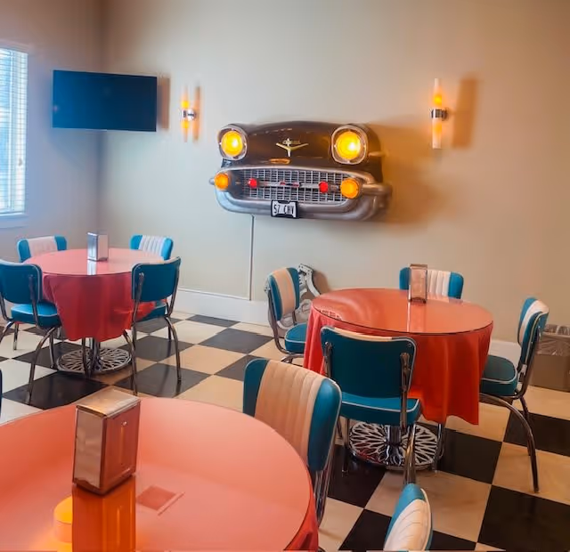 A retro-themed dining room with round tables covered in red tablecloths and teal and white vintage-style chairs. The floor has a black and white checkered pattern. On the wall, there is a decorative front end of a classic car with illuminated headlights, flanked by two wall sconces. A flat-screen TV is mounted on the adjacent wall near a window with blinds.