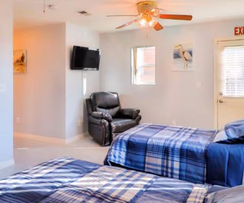 A bright bedroom with two beds covered in blue plaid bedding, a black leather recliner chair, a wall-mounted flat-screen TV, a ceiling fan with lights, and a door with a window and an exit sign above it. The walls are light-colored with a couple of framed pictures.
