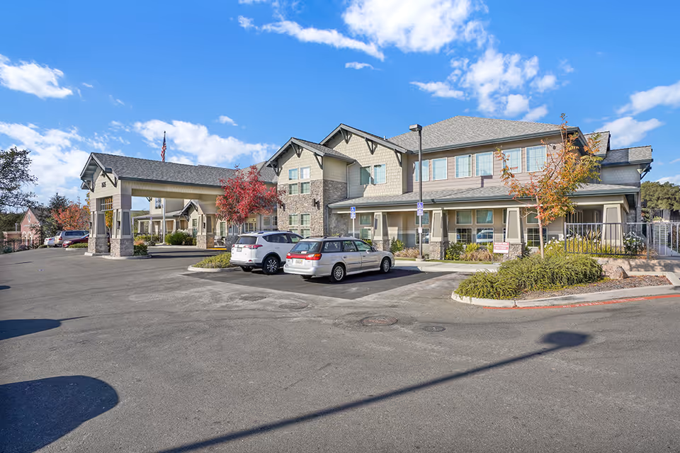 Exterior view of Almond Heights senior living facility showing a two-story building with a covered entrance, several parked cars, trees with autumn foliage, and a clear blue sky with scattered clouds.