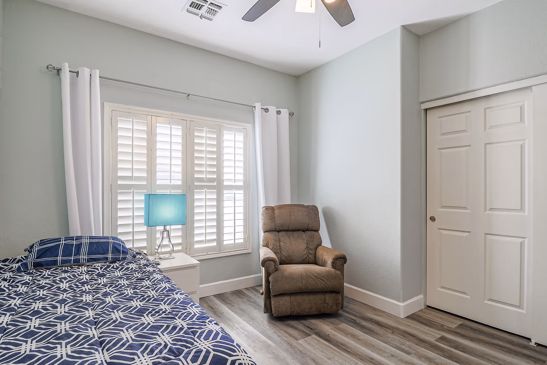 Bedroom with a patterned bed, white nightstand and blue lamp, a brown recliner, window shutters, and sliding closet doors.