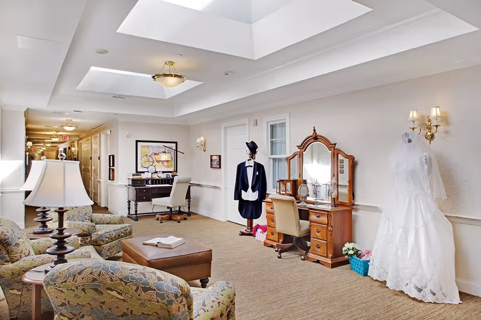 A well-lit interior common area with patterned armchairs, a leather ottoman with an open book, a wooden vanity with a mirror and chair, and a mannequin dressed in a white wedding gown. Another mannequin is dressed in a tuxedo. The space has beige walls, carpeted floors, ceiling lights, and skylights.