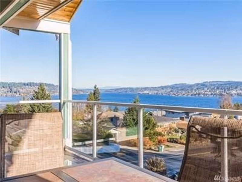 View from a balcony with a glass railing overlooking a body of water, distant hills, and residential houses. The balcony has a table and two wicker chairs under a wooden roof.