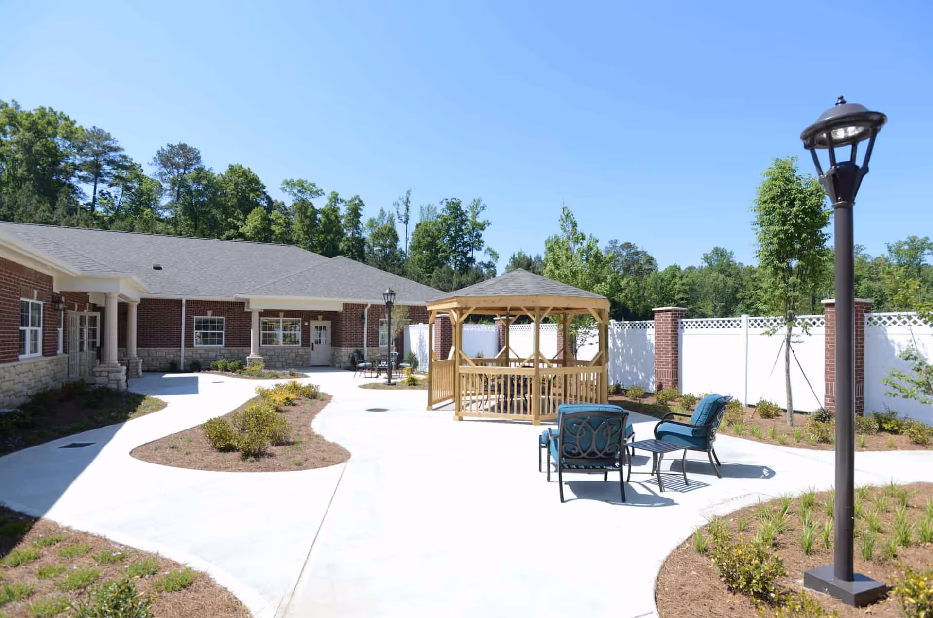 Outdoor courtyard area at the Reserve at Towne Lake featuring a wooden gazebo, patio chairs with cushions, a lamp post, landscaped garden beds, and a brick building with white trim in the background under a clear blue sky.