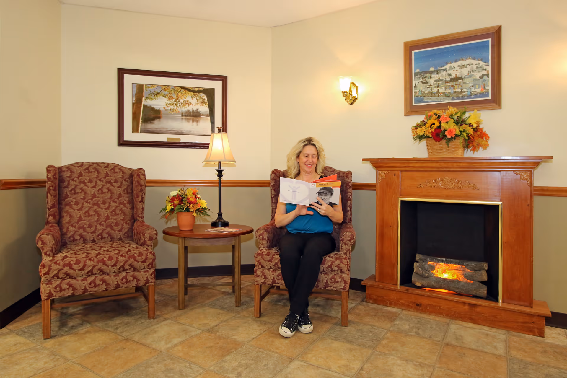 A woman sitting in a patterned armchair reading a brochure in a cozy room with a wooden fireplace, a side table with a lamp and a flower pot, and framed artwork on the walls.