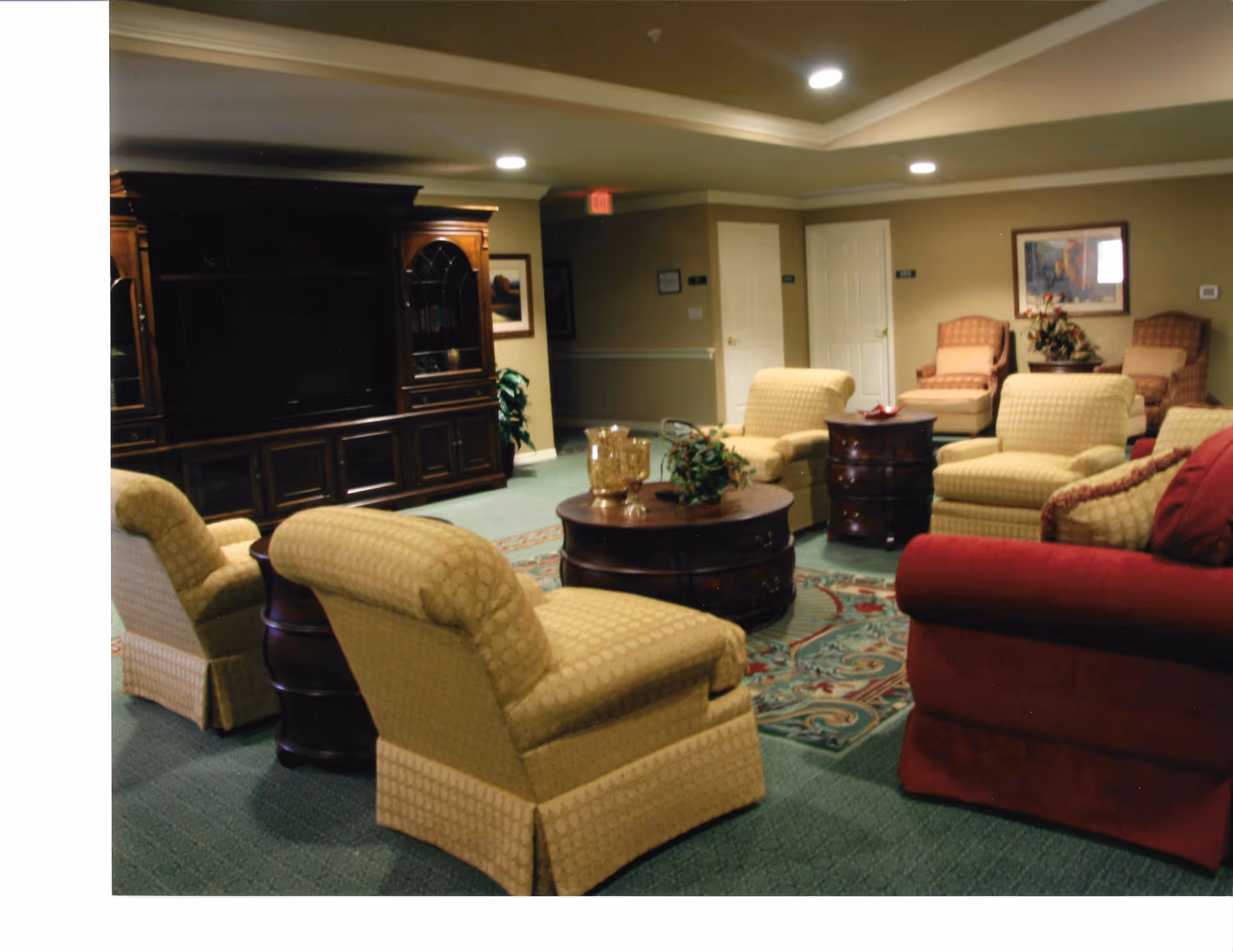 A cozy living room area with multiple upholstered armchairs in yellow and red, arranged around two wooden coffee tables on a patterned rug. There is a large dark wooden entertainment center against the wall, with framed artwork and a plant nearby. The room has soft lighting from recessed ceiling lights and beige walls with white trim.