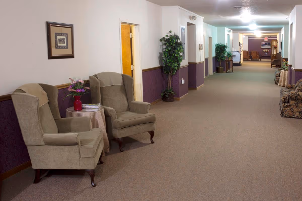 A long hallway in a senior living facility with beige carpet and walls painted white and purple. There are two beige armchairs with a small round table between them holding a vase with flowers and some magazines. The hallway has several doors, potted plants, framed pictures on the walls, and additional seating further down the corridor.