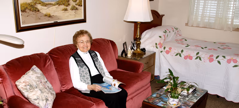 An elderly woman sitting on a red couch in a cozy room with a floral pillow, holding a magazine. Behind her is a bed with a white quilt decorated with pink flowers, a wooden nightstand with a lamp, and a window with white curtains. A coffee table with a plant and magazines is in front of the couch.