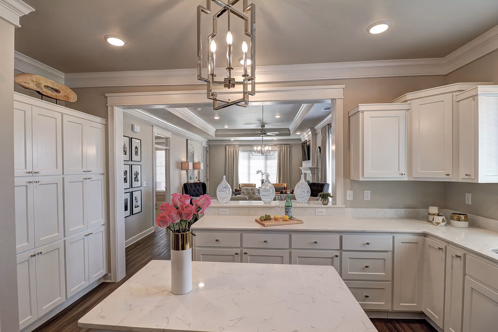 Modern kitchen with white cabinetry and marble countertops. A vase with pink flowers is on the kitchen island. In the background, a large mirror reflects a living room area with a chandelier, curtains, and furniture.