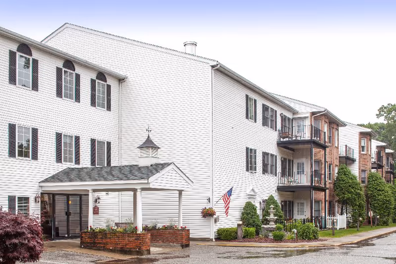 Exterior view of a multi-story senior living facility with white siding and brick accents. The building features multiple windows with black shutters, balconies, a covered entrance with a small cupola on top, and landscaped greenery including bushes and hanging flower baskets. An American flag is displayed near the entrance.