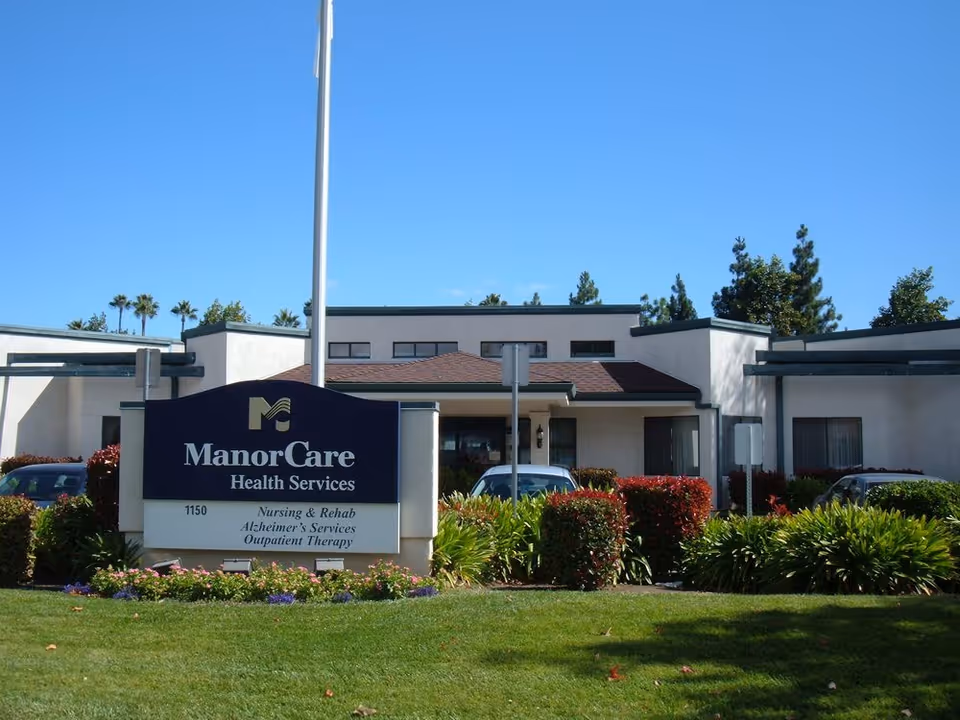 Front entrance of a single-story healthcare building with a large 'ManorCare Health Services' sign, landscaped lawn, and parked cars.