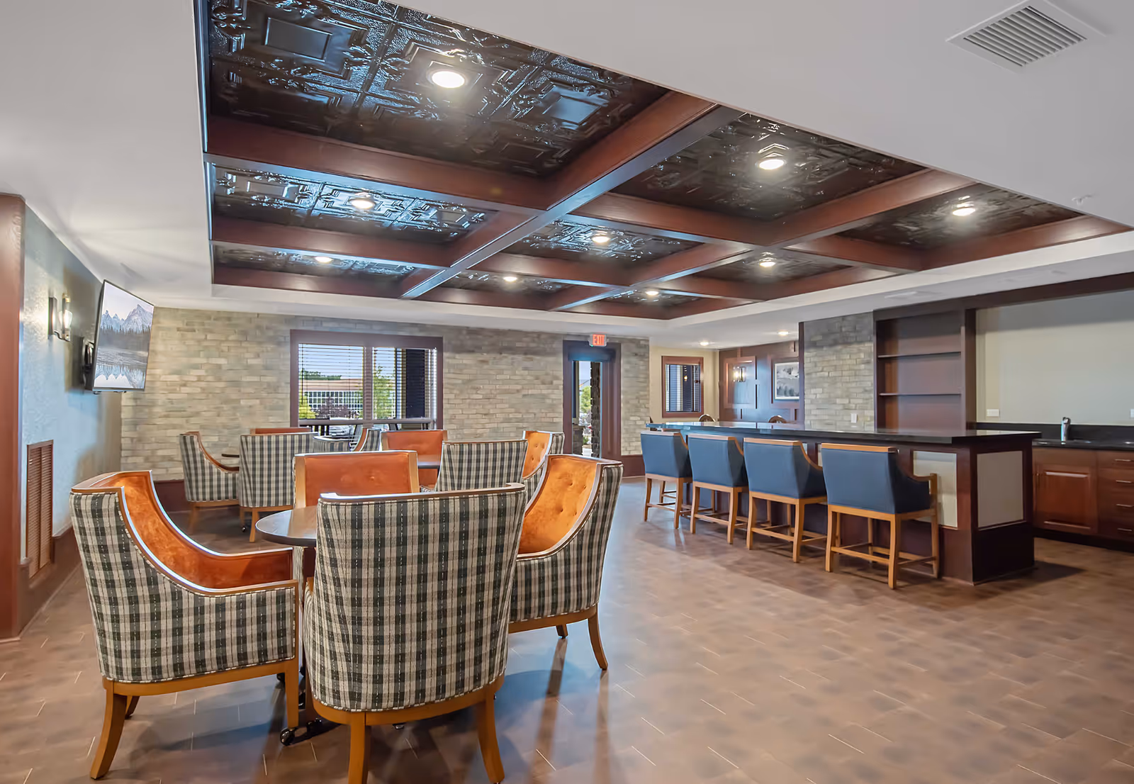 A spacious senior living common area with a checkered pattern of upholstered chairs around a round table in the foreground and a long bar counter with blue cushioned bar stools in the background. The room features a decorative coffered ceiling with recessed lighting, brick accent walls, a wall-mounted TV, and large windows letting in natural light.