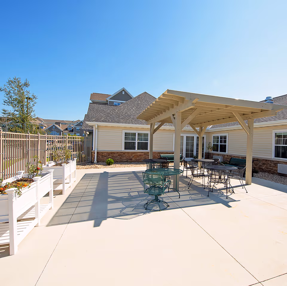 Outdoor patio area at All Saints Memory Care with a pergola providing shade over several metal tables and chairs. Raised garden beds with plants line one side, and the building with beige siding and stone accents is visible in the background under a clear blue sky.