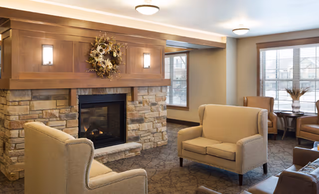 A cozy living room area with a stone fireplace decorated with a floral wreath above the mantle. The room features beige upholstered armchairs and a loveseat arranged around the fireplace, with large windows letting in natural light and a small table with a vase in the corner.