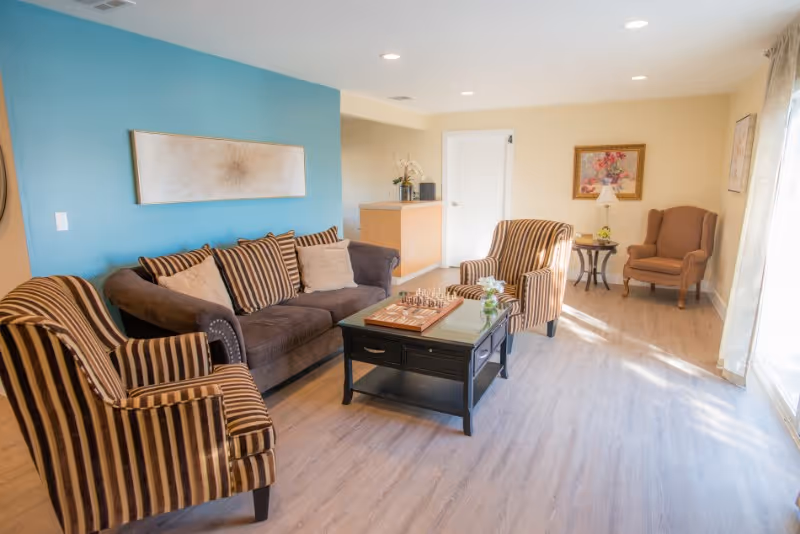 A bright living room with a brown couch and two striped armchairs around a black coffee table with a chess set on it. The room has light wood flooring, a blue accent wall with a rectangular piece of artwork, and a beige wall with a framed floral painting and a small table with a lamp. Sunlight streams in through a large window with sheer curtains.