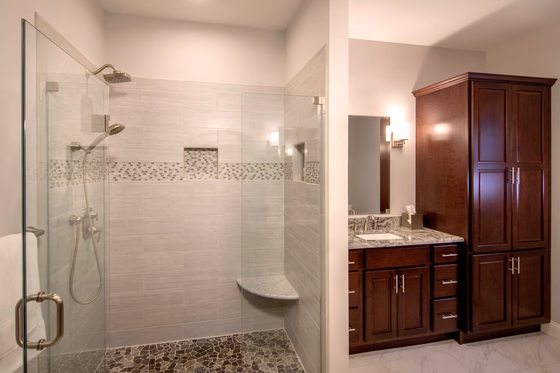 Modern bathroom with a glass-enclosed walk-in shower featuring a built-in bench, dual shower heads, and decorative tile accents. Adjacent to the shower is a vanity with a granite countertop, a sink, a mirror, wall-mounted light fixtures, and dark wood cabinetry including a tall storage cabinet.