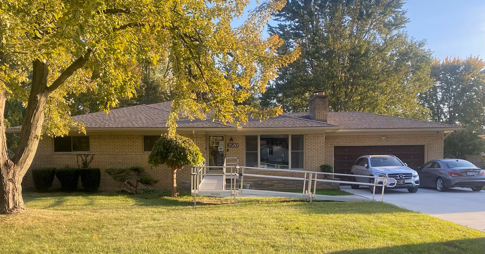 Single-story brick building with a sloped roof, a wheelchair ramp leading to the front door, two cars parked in the driveway, and trees with green and yellow leaves surrounding the property.