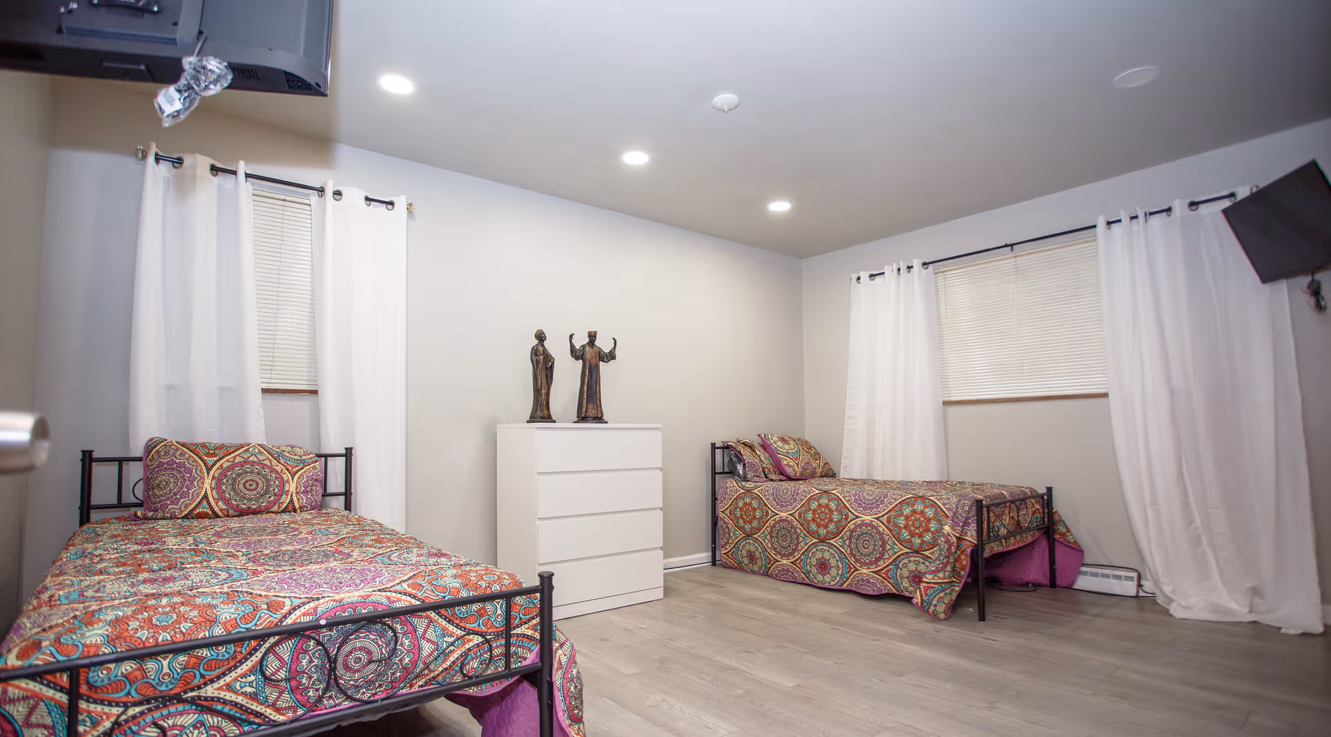 A bright shared bedroom with two twin beds, colorful patterned bedspreads, a white dresser and white curtains.
