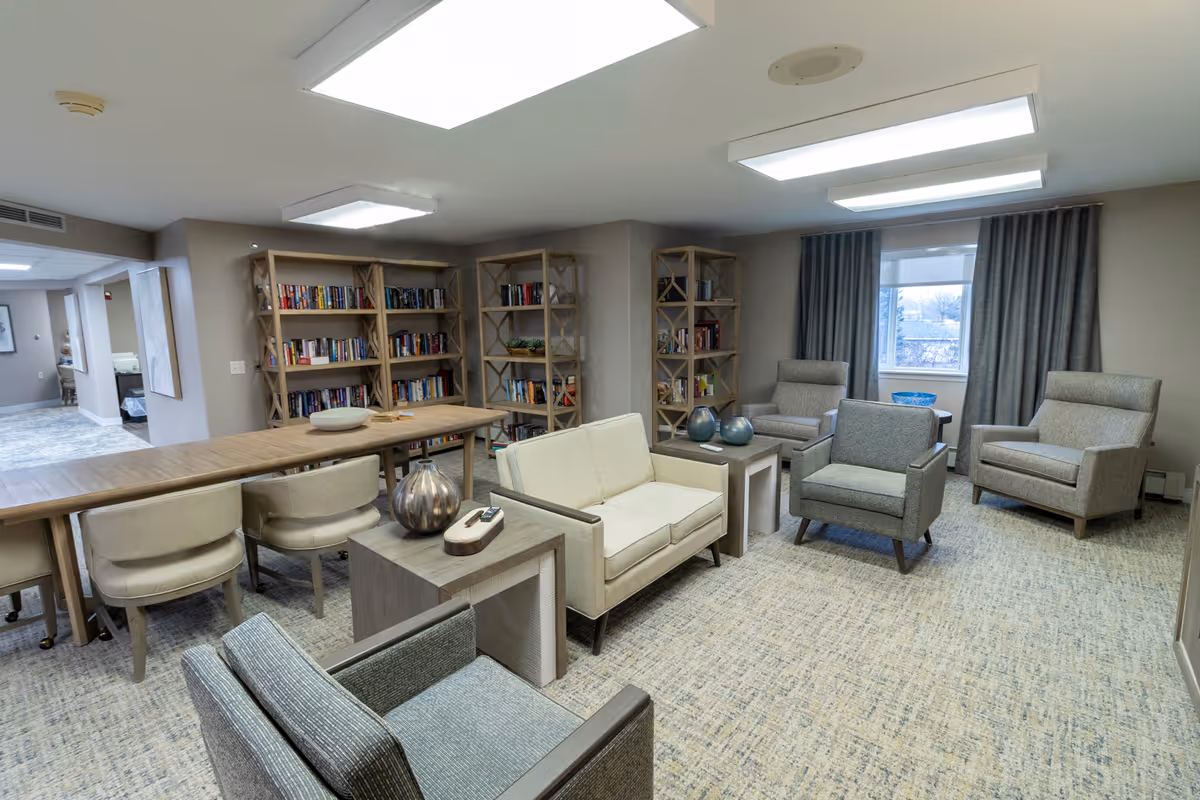 A cozy common area in a senior living facility featuring multiple armchairs and a loveseat arranged around small side tables with decorative vases. Behind the seating area are wooden bookshelves filled with books. A long table with chairs is positioned near the bookshelves. The room has large ceiling lights and a window with gray curtains letting in natural light.