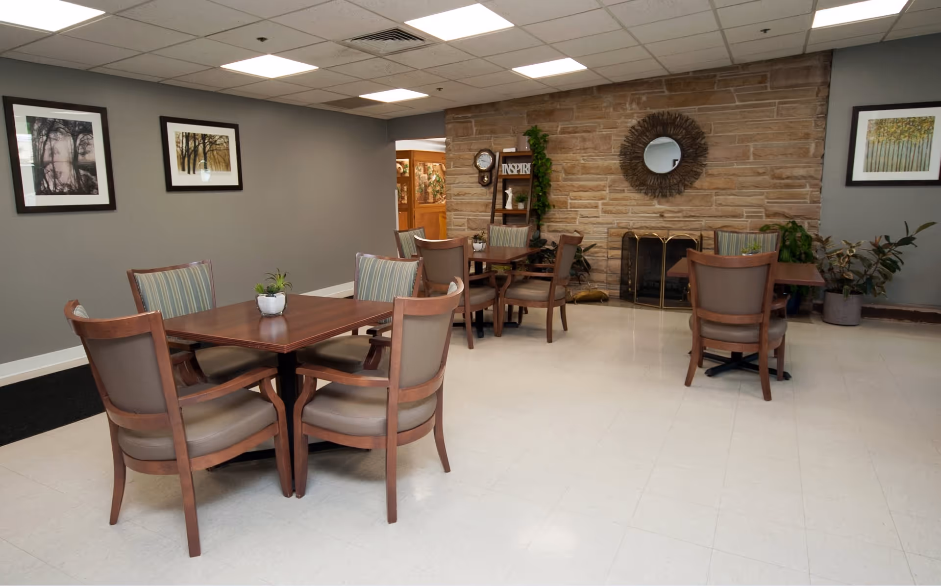 A senior living facility common area with several wooden tables and cushioned chairs arranged around them. The room has a stone fireplace with a decorative round mirror above it, a wall clock, and a small shelf with plants and decor. The walls are painted gray and adorned with framed nature artwork. The floor is light-colored tile, and there are potted plants near the fireplace.