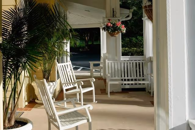 A covered outdoor porch area with white wooden rocking chairs and a white wooden bench. There is a hanging basket with pink flowers and a large potted plant on the left side. The porch overlooks a parking area and greenery in the background.