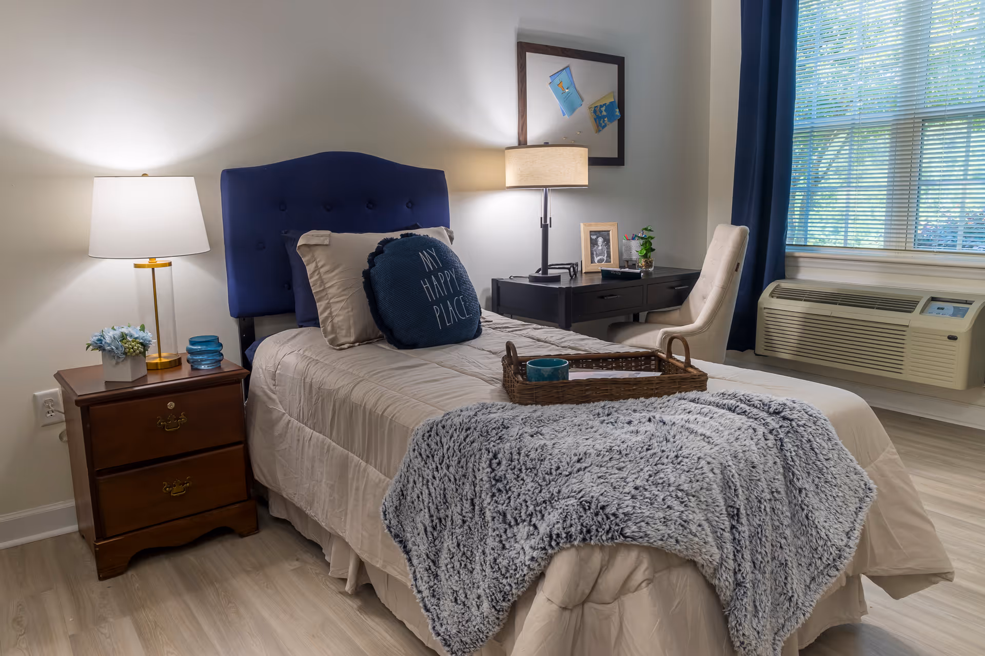 A cozy bedroom with a single bed featuring a blue upholstered headboard, beige bedding, and a decorative pillow that says 'MY HAPPY PLACE'. A wooden nightstand with a lamp, small flower arrangement, and blue glass decor is beside the bed. Across from the bed is a black desk with a beige chair, a table lamp, a framed photo, and a small plant. A window with blue curtains and an air conditioning unit is on the right side of the room.