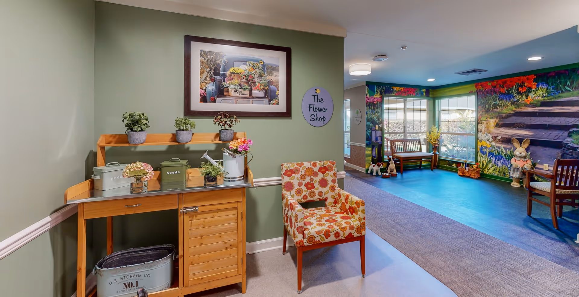 Interior view of a senior living facility area with a green wall featuring a framed picture of a truck with flowers and a sign that reads 'The Flower Shop'. A wooden cabinet with potted plants, seed containers, and a watering can is next to a floral-patterned armchair. The adjacent room has a large mural of a garden with flowers and stairs, a wooden bench, and stuffed animal decorations.