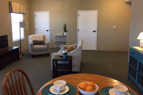 Small living/dining area with a round table set with cups and a bowl of oranges in the foreground, armchairs and a sofa facing a TV stand, and two white doors on a beige wall.