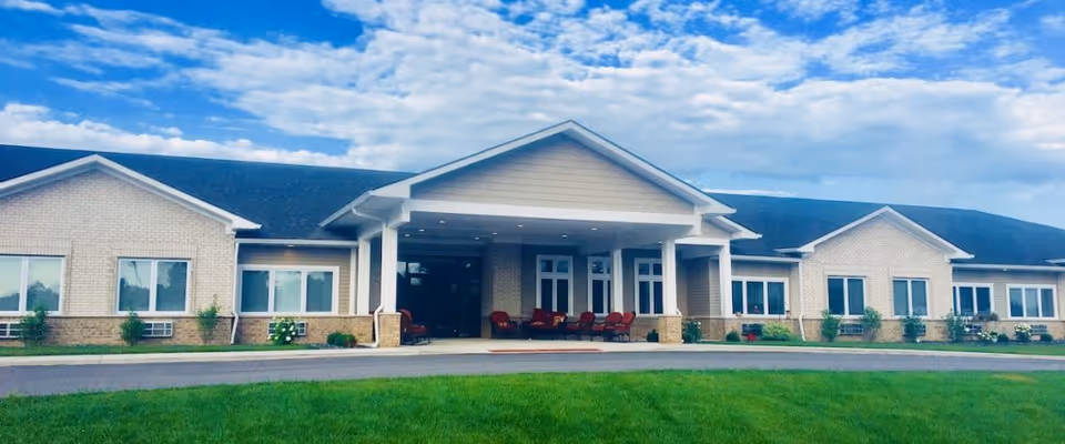 Front exterior of a single-story brick assisted living building with a covered entrance, chairs on the porch, and a manicured lawn under a blue sky.