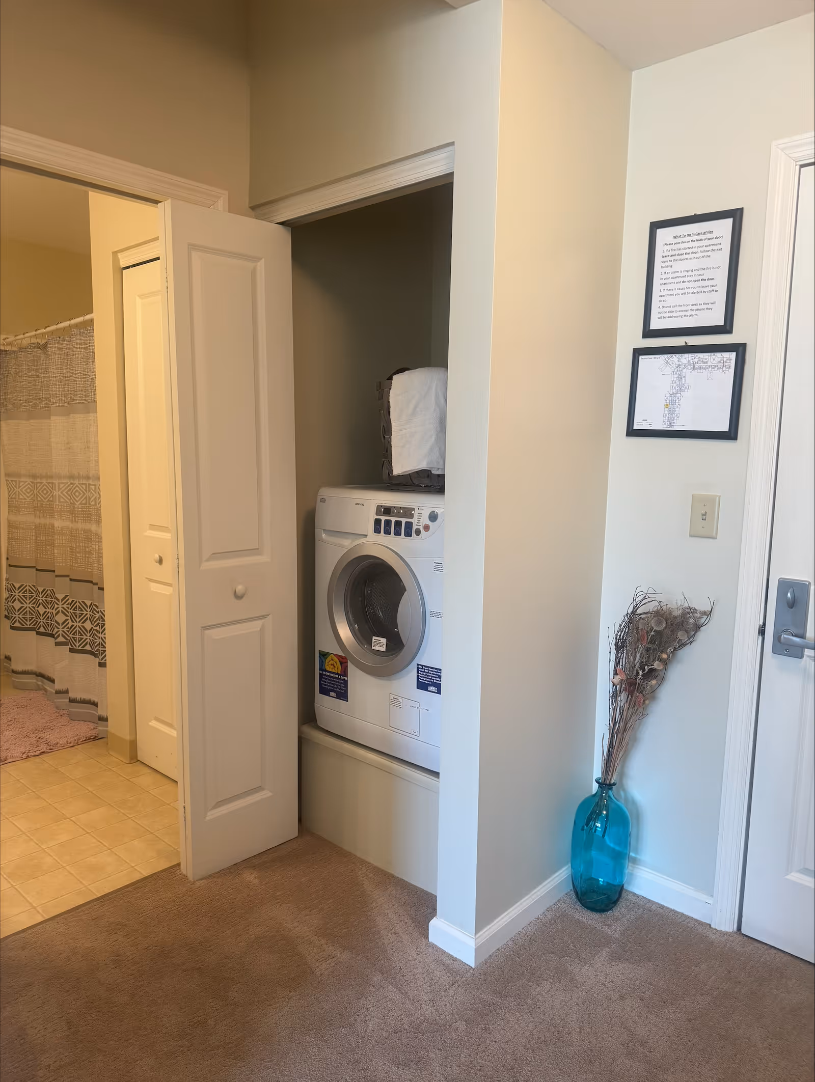 Interior hallway corner with a stacked washer/dryer in a closet, an open bathroom door with a shower curtain, and a blue vase holding decorative branches.