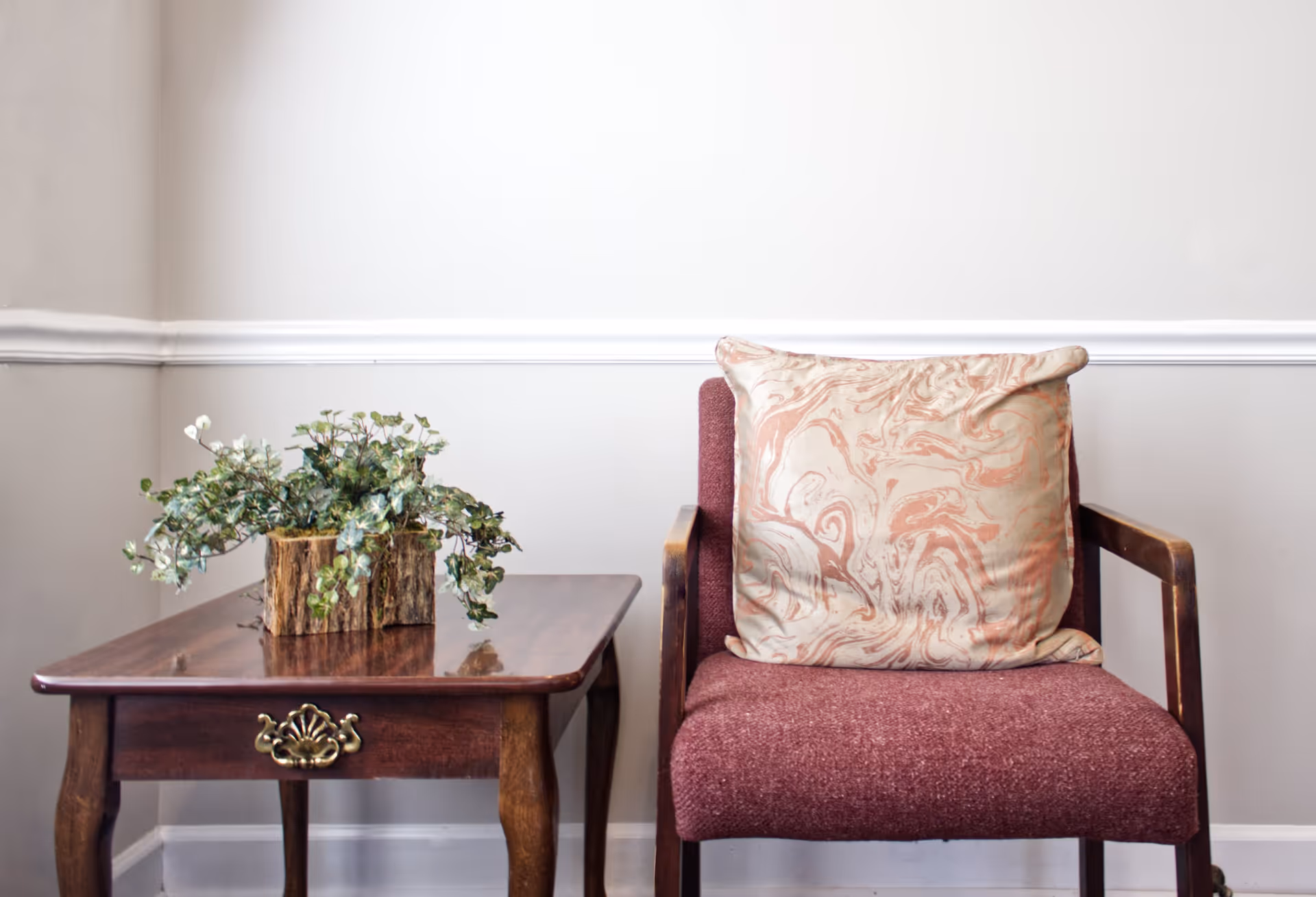 A cozy corner featuring a wooden side table with a decorative plant in a rustic wooden planter, next to a maroon upholstered chair with wooden armrests and a large cushion with a pink and beige marbled pattern, set against a light gray wall with white trim.