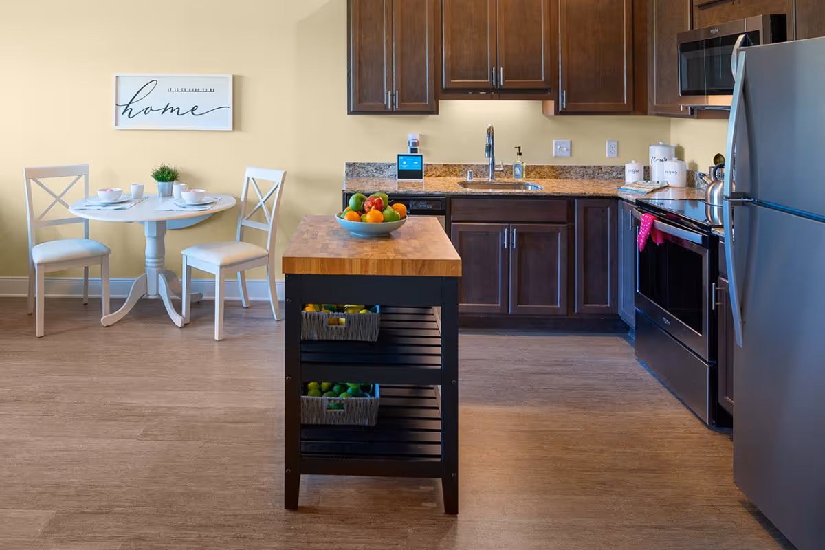 A modern kitchen with dark wood cabinets, granite countertops, stainless steel refrigerator and oven, a small kitchen island with baskets of fruit, and a round white dining table with two chairs set with bowls and cups. A wall decoration with the word 'home' is hung above the dining table.