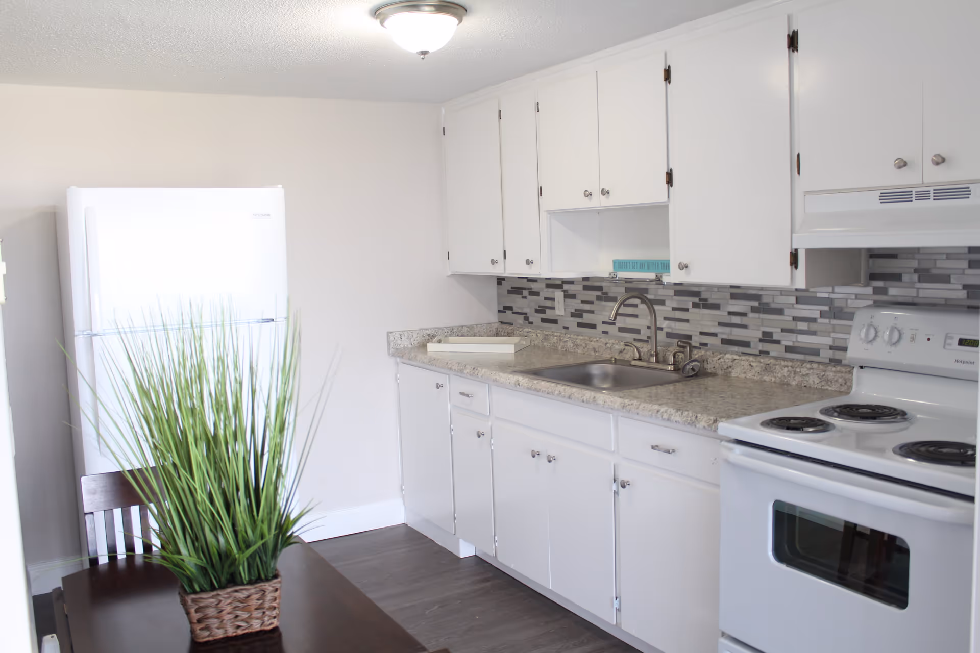A bright kitchen with white cabinets, a white refrigerator, a white electric stove, a stainless steel sink, and a countertop with a gray and white mosaic tile backsplash. A dark wooden table with a green potted plant is partially visible in the foreground.