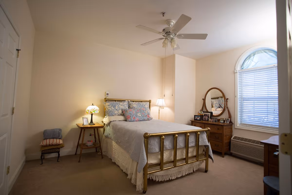 Well-lit bedroom with a brass bed, bedside tables, dresser with mirror, and an arched window.