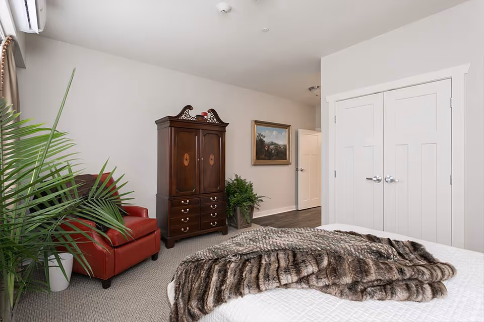 A bedroom with a white bed covered by a brown and gray fur throw blanket. To the left, there is a red armchair next to a large green potted plant. Against the far wall, there is a dark wooden armoire with drawers and decorative details on top. A framed painting hangs on the wall near a hallway with an open door. On the right side, there is a white double-door closet.
