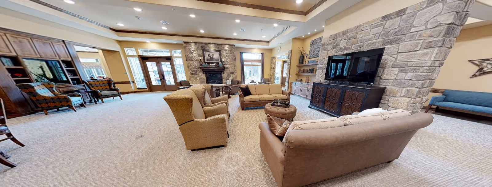 A spacious and well-lit living room area in a senior living facility featuring multiple comfortable armchairs and sofas arranged around a central ottoman. The room has a stone fireplace with a framed picture above it, large windows allowing natural light, a flat-screen TV mounted on a stone wall, wooden cabinetry, and decorative shelves. The carpeted floor and warm color tones create a cozy and inviting atmosphere.